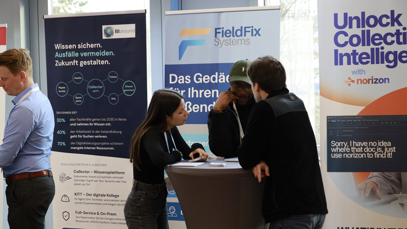 Drei Personen unterhalten sich an einem Stehtisch. Im Hintergrund sind Roll-ups verschiedener Start-ups zu sehen.