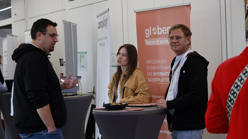 Drei Personen, zwei Männer und eine Frau, stehen an einem Stehtisch und unterhalten sich.